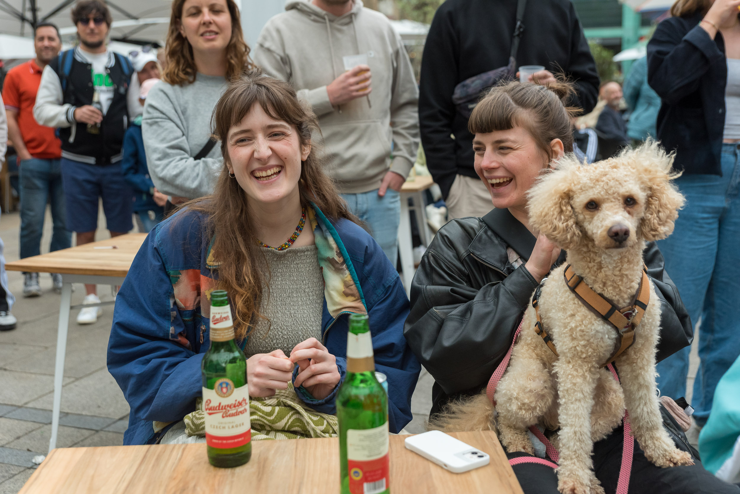 Zwei Junge Frauen sitzen auf Bänken in einer Zuschauerreihe und lachen, eine hat einen Hund auf dem Schoß.